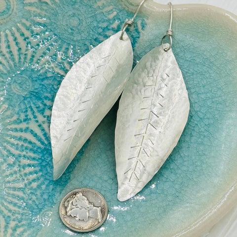 JANECKA Sterling Silver Hand Forged Leaf Earrings by Janecka, featuring etched vein details, are shown on a blue ceramic dish beside a U.S. quarter for scale.