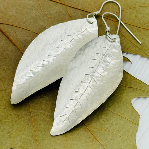 The Janecka JANECKA Sterling Silver Hand Forged Leaf Earrings feature a hammered texture and stitched detailing, shown atop a dried leaf.