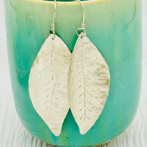JANECKA Sterling Silver Hand Forged Leaf Earrings by Janecka feature textured surfaces and delicate etched veins, displayed against a glossy turquoise and green ceramic backdrop.