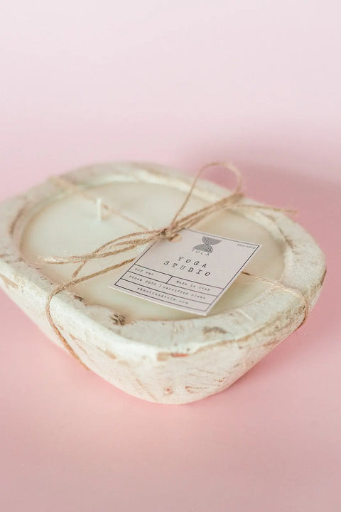 The Shanti + Tula Hand-Poured Soy Dough Bowl Candle, in rustic or antique white, features three wicks in an oval bowl, tied with twine and a Yoga Studio tag, adding a warm touch against a soft pink background. Made in the USA.