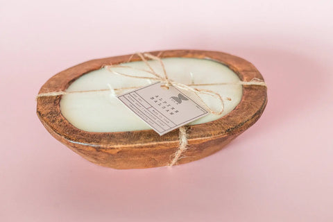 The Shanti + Tula Hand-Poured Soy Dough Bowl Candle – Oval 3-Wick, in Rustic or Antique White, adds charm to any space. Made in the USA and crafted from 100% soy wax, its pictured with a product tag against a pale pink background.