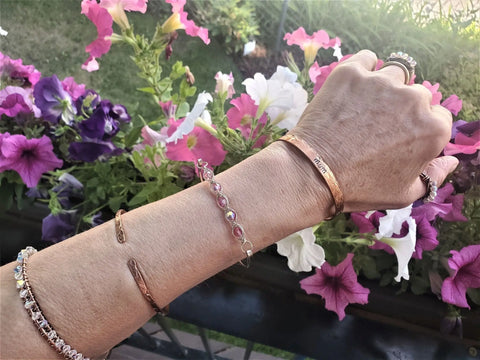 A hand and forearm wear the Alexa Martha Designs MOM Stamped Copper Cuff, extended over a balcony railing with blooming pink, purple, and white petunias and green grass in the background.