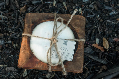 The Shanti + Tula Handcrafted 3-Wick Square Dough Bowl Candle, made with soy wax and finished in natural & antique white, sits on a rustic wooden tray with a Palo Santo tag amid dark mulch and leaves outdoors. Made in the USA.