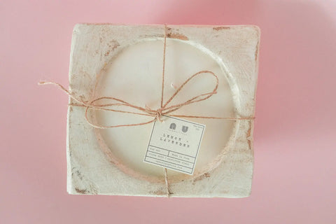 The Shanti + Tula Handcrafted 3-Wick Square Dough Bowl Candle, made from soy wax with a natural & antique white finish, exudes eco-conscious style and charm against a soft pink background. Made in the USA.