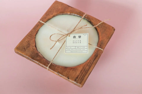 The Shanti + Tula Handcrafted 3-Wick Square Dough Bowl Candle, made in the USA with soy wax and a natural & antique white finish, is displayed against a light pink background.