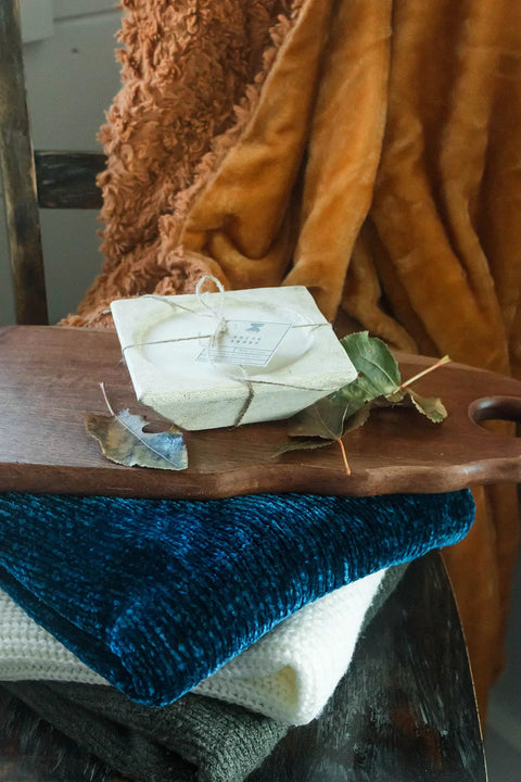 A Shanti + Tula Handcrafted 3-Wick Square Dough Bowl Candle in natural and antique white rests on folded blue, white, and gray textured blankets, with a fuzzy orange blanket draped softly in the background.