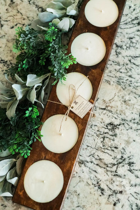 The Shanti + Tula Handcrafted 5-Hole Cheese Mold Candle is a 28 soy dough bowl centerpiece, featuring five hand-poured white candles in a wooden tray, accented with a tag and displayed beside artificial green and silver foliage.