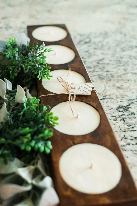 The Shanti + Tula Handcrafted 5-Hole Cheese Mold Candle—a 28 soy dough bowl centerpiece—features five white candles with twine and tag, displayed on a textured light surface with green leafy plants beside it. Made in the USA.