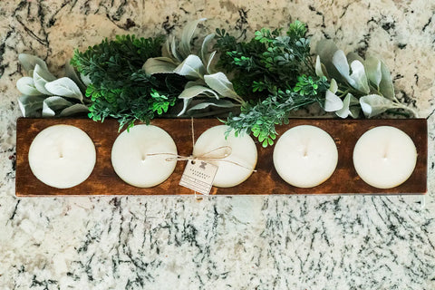 The Shanti + Tula Handcrafted 5-Hole Cheese Mold Candle is a 28 soy dough bowl centerpiece featuring five white candles with green leafy accents and a small brown tag, beautifully displayed on a light marbled surface.