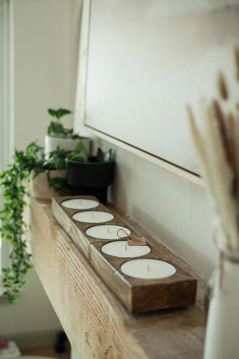 A close-up of the Shanti + Tula Handcrafted 5-Hole Cheese Mold Candle—a 28 soy dough bowl centerpiece—sits on a wooden shelf with potted plants and dried grasses, creating a cozy, natural decor arrangement.