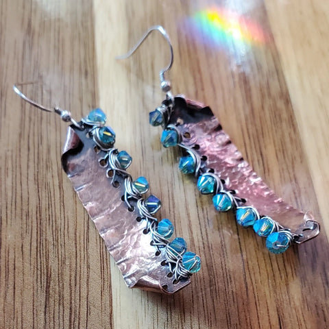 The Blue Green Hammered Copper Bar Crystal Earrings by Alexa Martha Designs feature a textured finish, iridescent blue beads, and silver wire wrapping, displayed on wood with a subtle rainbow reflection nearby.