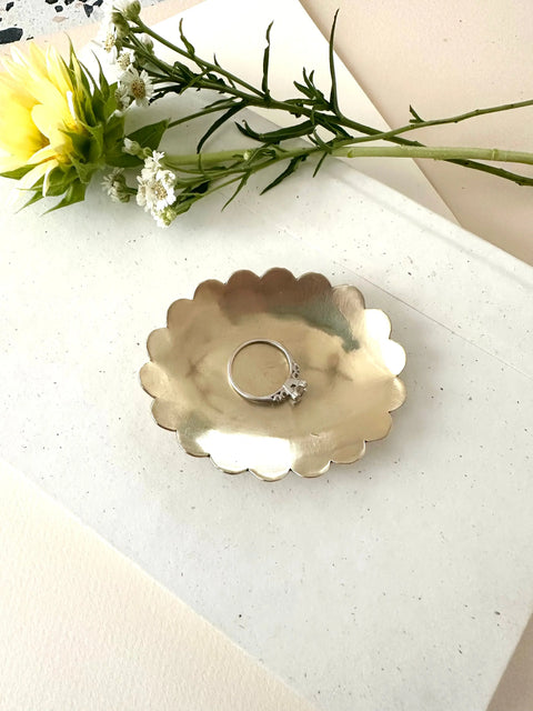 The BRASS SCALA JEWELRY DISH by Sarah Cecelia, featuring scalloped edges, elegantly displays a gemstone silver ring on a white textured surface with a yellow flower and greenery—an ideal engagement gift.
