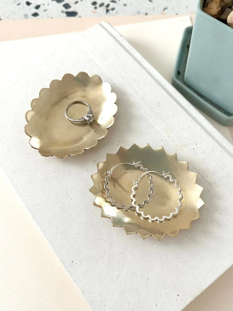 Two Sarah Cecelia BRASS SCALA JEWELRY DISHES rest on a white surface—one with a silver ring, the other with wavy hoop earrings. A blue planter with pebbles appears nearby, making this set a charming engagement gift.