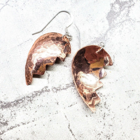 The Broken Heart Earrings From Sculpted Copper by Alexa Martha Designs feature hammered, jagged-edged halves of a heart, artfully displayed on a white and gray marbled background for a distinctive look.