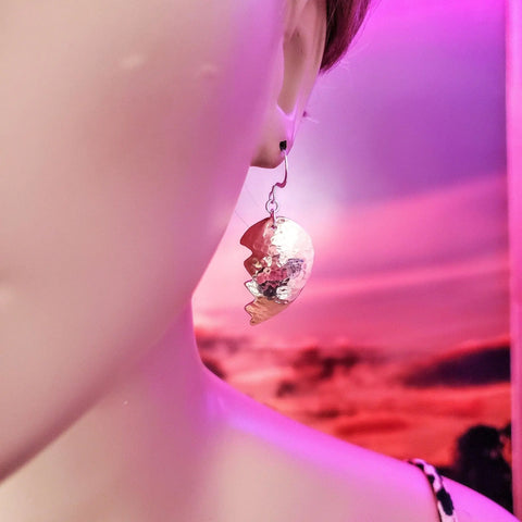 A close-up shows a person wearing Broken Heart Earrings From Sculpted Copper by Alexa Martha Designs. The dreamy, pink and purple sky background highlights the earrings’ unique design.