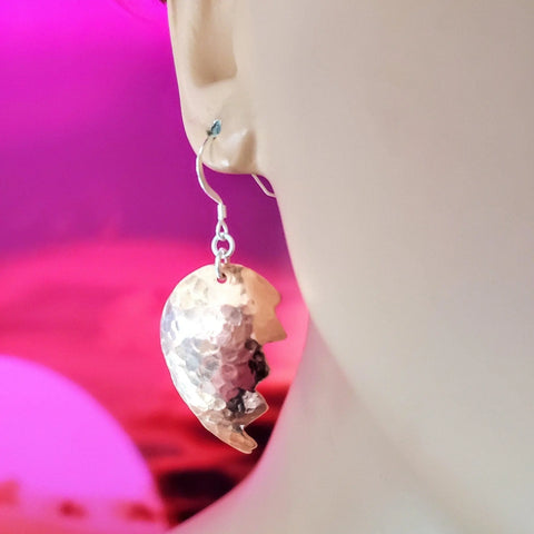 A close-up of a person wearing Broken Heart Earrings from Sculpted Copper by Alexa Martha Designs, shown against a vibrant pink and purple background.