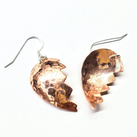 The Broken Heart Earrings from Sculpted Copper by Alexa Martha Designs feature asymmetrical, jagged-edge halves with a hammered texture, highlighting their unique design against a white background.