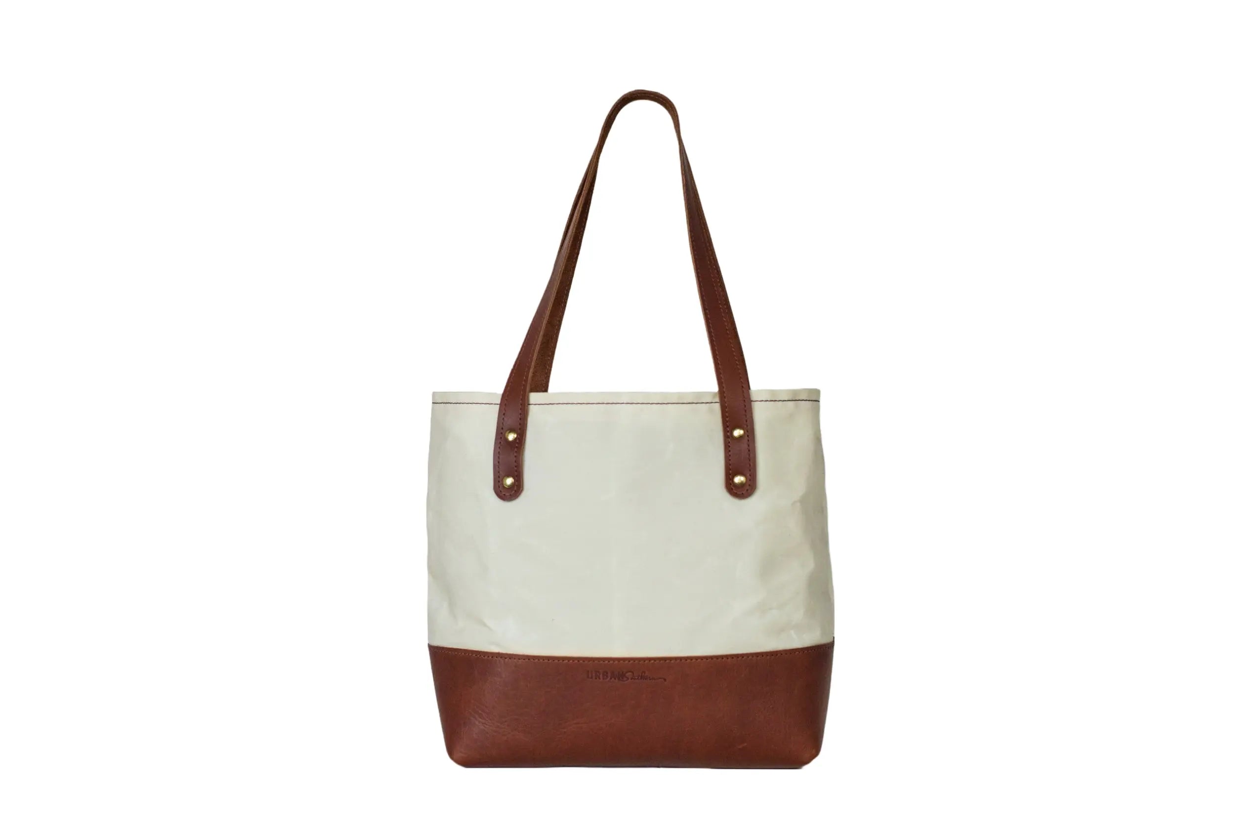 The Urban Southern Classic Canvas Tote features a white canvas upper, brown leather bottom, two brown leather straps, and gold-tone rivets—crafted in the USA.