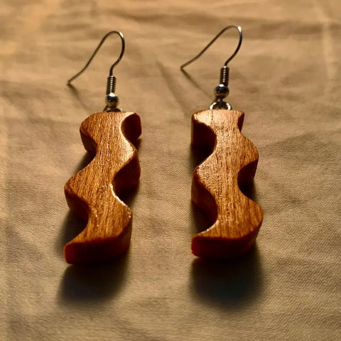 The Squiggle Earring by Shop Cat Woodworks is a pair of handcrafted wooden earrings with a wavy design, shown on beige fabric under warm light that highlights their rich brown color and natural wood grain—an elegant, eco-conscious jewelry choice.