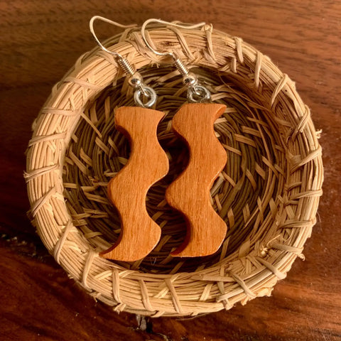 The Squiggle Earring by Shop Cat Woodworks is a handcrafted wooden pair with wavy, abstract shapes and silver hooks, elegantly presented in a small woven basket—an eco-conscious jewelry piece.