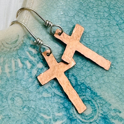 JANECKA Copper Cross Earrings by Janecka, hand-forged with sterling silver ear wires, shown on a textured blue ceramic surface.
