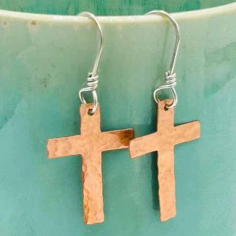 The JANECKA Copper Cross Earrings by Janecka, hand forged with sterling silver ear wires, are shown hanging against a turquoise background.