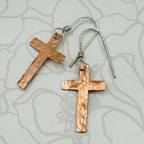 The JANECKA Copper Cross Earrings by Janecka are hand forged and feature sterling silver ear wires, displayed on a light gray patterned background—unique handmade jewelry for any occasion.