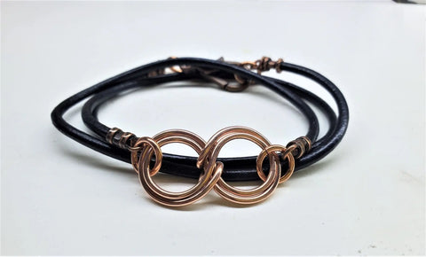 The Copper Double Infinity Gender Neutral Leather Wrap Bracelet by Alexa Martha Designs features a central copper double infinity wire on black leather, elegantly displayed on a white surface.