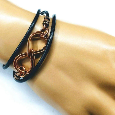 A hand displays the Alexa Martha Designs Copper Double Infinity Gender Neutral Leather Wrap Bracelet, featuring black leather with a copper wire double infinity symbol centerpiece.