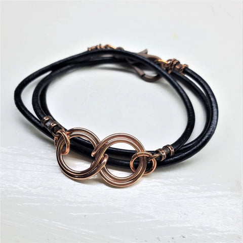 The Copper Double Infinity Gender Neutral Leather Wrap Bracelet by Alexa Martha Designs features two interlocking copper rings on black leather—an elegant accessory for any style, shown on a white surface.
