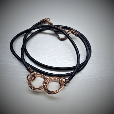 The Copper Double Infinity Gender Neutral Leather Wrap Bracelet by Alexa Martha Designs features black leather with two interlocking metallic rings and clasp, resembling a double infinity, displayed on a white surface.