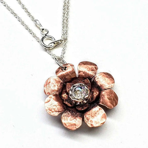 The Copper Crystal Flower Blossom Spinner Necklace by Alexa Martha Designs features a hammered copper flower pendant with layered petals and a clear gemstone center on a silver chain, offering elegant fidget style. Displayed on a white background.