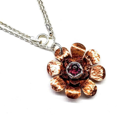 The Copper Crystal Flower Blossom Spinner Necklace by Alexa Martha Designs features hammered copper petals, a central red gemstone, and a silver chain. This handmade Montana fidget necklace is shown on a white background.
