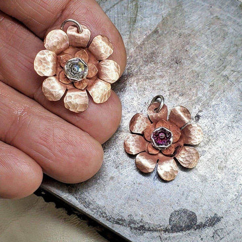 A hand holds one of two hammered Copper Crystal Flower Blossom Spinner Necklaces by Alexa Martha Designs—silver-colored and purple—with gemstone centers, handmade in Montana for unique, calming style.