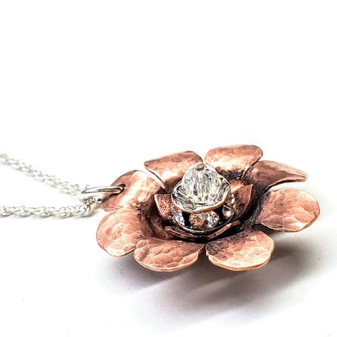 A close-up of the Copper Crystal Flower Blossom Spinner Necklace by Alexa Martha Designs, handmade in Montana with textured copper petals and a clear gemstone center, displayed on a silver chain against a white background.