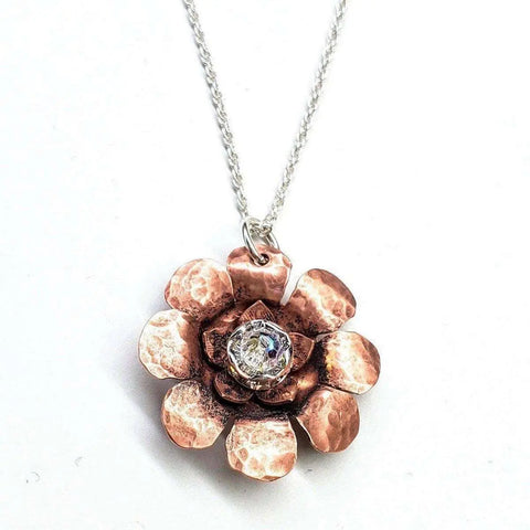 The Copper Crystal Flower Blossom Spinner Necklace by Alexa Martha Designs features a hammered copper flower pendant with layered petals and a sparkling clear gem, handmade in Montana and shown on a white background.