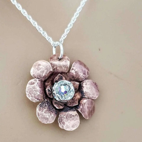 A close-up of the Alexa Martha Designs Copper Crystal Flower Blossom Spinner Necklace, featuring a textured metallic flower with a clear crystal center—an elegant fidget necklace for subtle, soothing motion against a beige background.