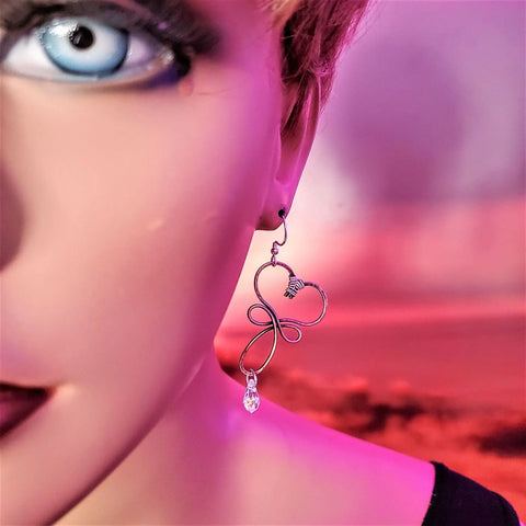 Close-up of a person’s face with a blue eye and dark makeup, wearing Alexa Martha Designs’ Crystal Drop Copper Heart Earrings. Each heart features a dangling Swarovski crystal. The background is blurred in pink and red tones.