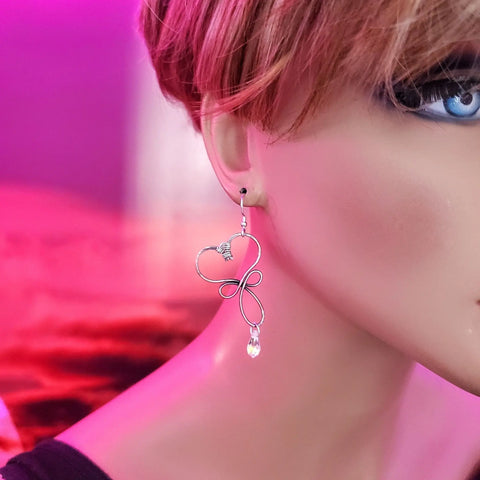 Close-up of a mannequin wearing Alexa Martha Designs Crystal Drop Copper Heart Earrings, featuring a large heart shape with a dangling bead, set against a pink and purple background. Only part of the face and ear are shown.