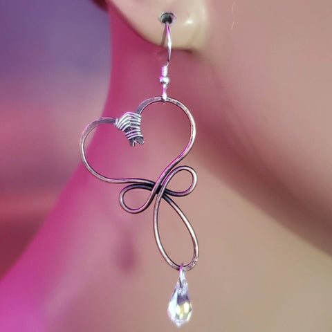 A close-up of a person’s ear wearing Alexa Martha Designs Crystal Drop Copper Heart Earrings, handmade with copper wire and a dangling Swarovski crystal teardrop, set against a soft pink and purple background.