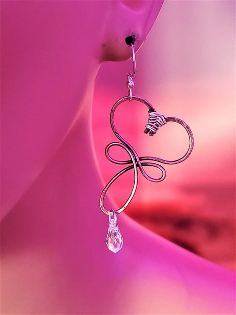A close-up of a person wearing Alexa Martha Designs’ Crystal Drop Copper Heart Earrings, featuring a teardrop crystal pendant, against a pink and purple background.