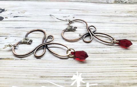 The Crystal Drop Copper Heart Earrings by Alexa Martha Designs feature handcrafted copper wire hearts, silver accents, and faceted red Swarovski crystal drops—an ideal gift for handmade copper jewelry lovers. Displayed on a light wooden surface.