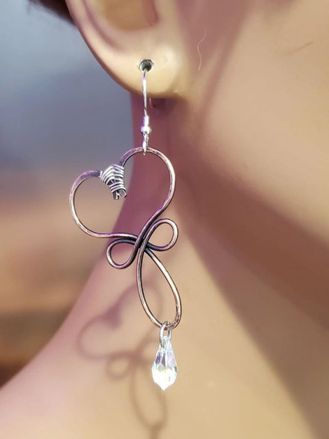 A close-up of a person wearing Crystal Drop Copper Heart Earrings by Alexa Martha Designs, featuring handmade wire wrapping, a swirl heart shape, and a dangling clear teardrop bead—ideal for fans of unique copper jewelry.