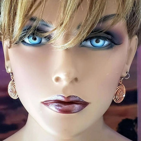 Close-up of a mannequin’s face with striking blue eyes, dramatic makeup, short blond hair, and wearing Alexa Martha Designs Crystal Embedded Copper Spiral Hoop Earrings against a blurry purple and dark-toned background.