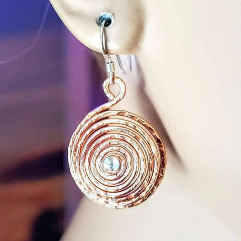 A close-up of Alexa Martha Designs Crystal Embedded Copper Spiral Hoop Earring, featuring gold-toned concentric circles and a central blue Swarovski Crystal, shown worn on an ear.
