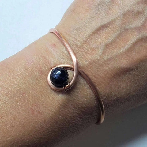 A close-up of a wrist wearing the Handmade Copper Ribbon Child Abuse Awareness Teardrop Bangle by Alexa Martha Designs, featuring a black faceted center stone and minimalist design with an elegant crackle agate bead.
