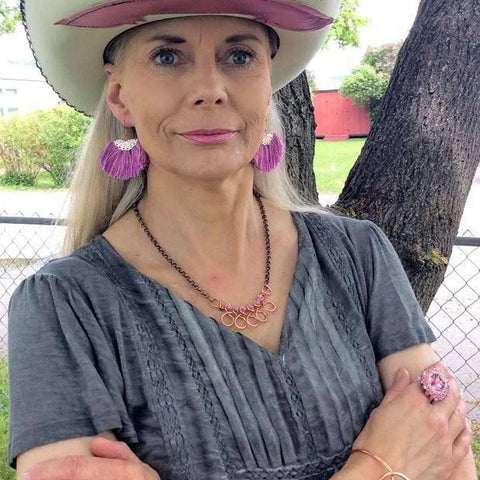 A woman with long blonde hair, a white cowboy hat, and pink earrings stands outdoors by a tree and fence, wearing handcrafted jewelry and the Handmade Copper Teardrop Bangle by Alexa Martha Designs.