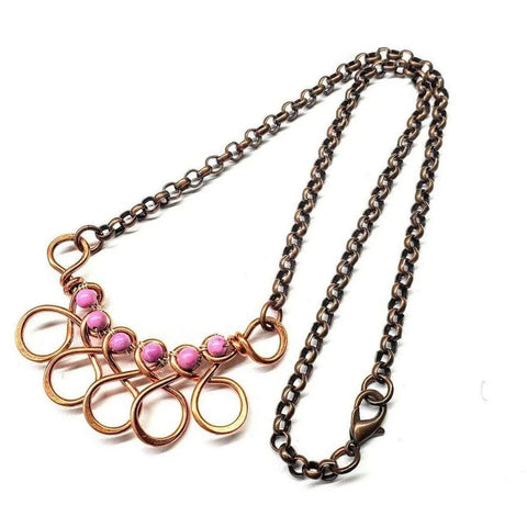 The Copper Wire Wrapped Sculpted Pink Gemstone Necklace by Alexa Martha Designs features loops and swirls with five pink beads, a bronze chain, lobster clasp closure, and is displayed against a white background.