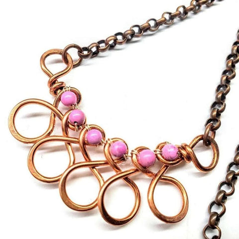 The Copper Wire Wrapped Sculpted Pink Gemstone Necklace by Alexa Martha Designs features handmade copper wire loops with five pink gemstones on a dark metal chain, creating a unique artisan statement piece.
