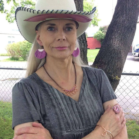 A woman in a gray dress and white cowboy hat stands outdoors with her arms crossed, wearing pink earrings, a pink ring, and the Alexa Martha Designs Copper Wire Wrapped Sculpted Pink Gemstone Necklace.